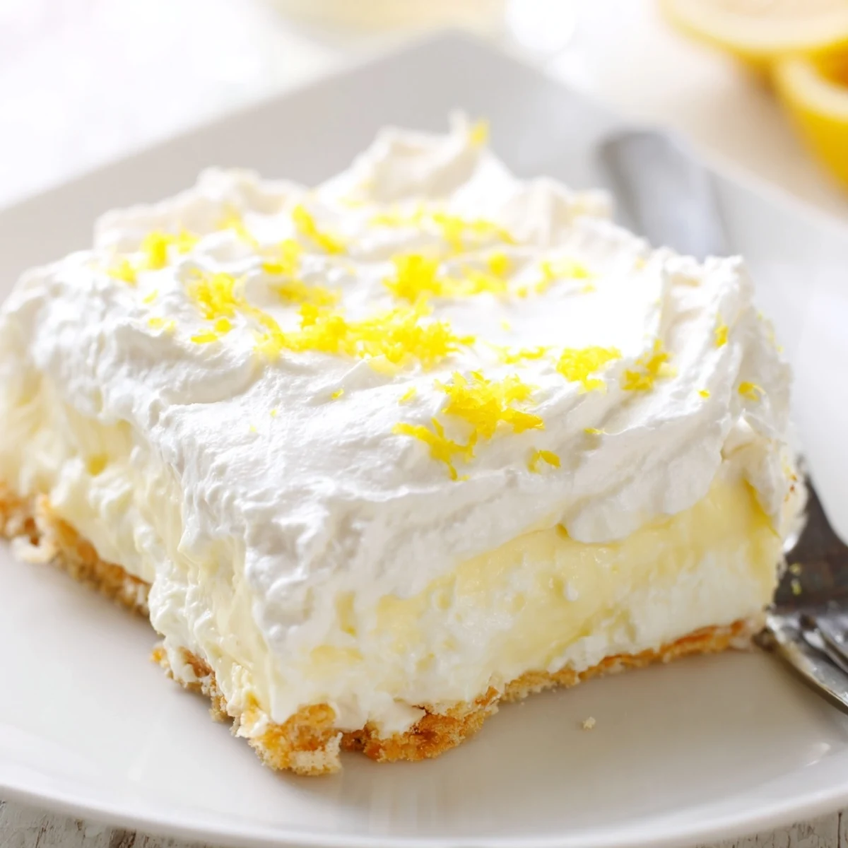 Square slice of chilled Lemon Lush Cake topped with bright lemon zest on a white dessert plate